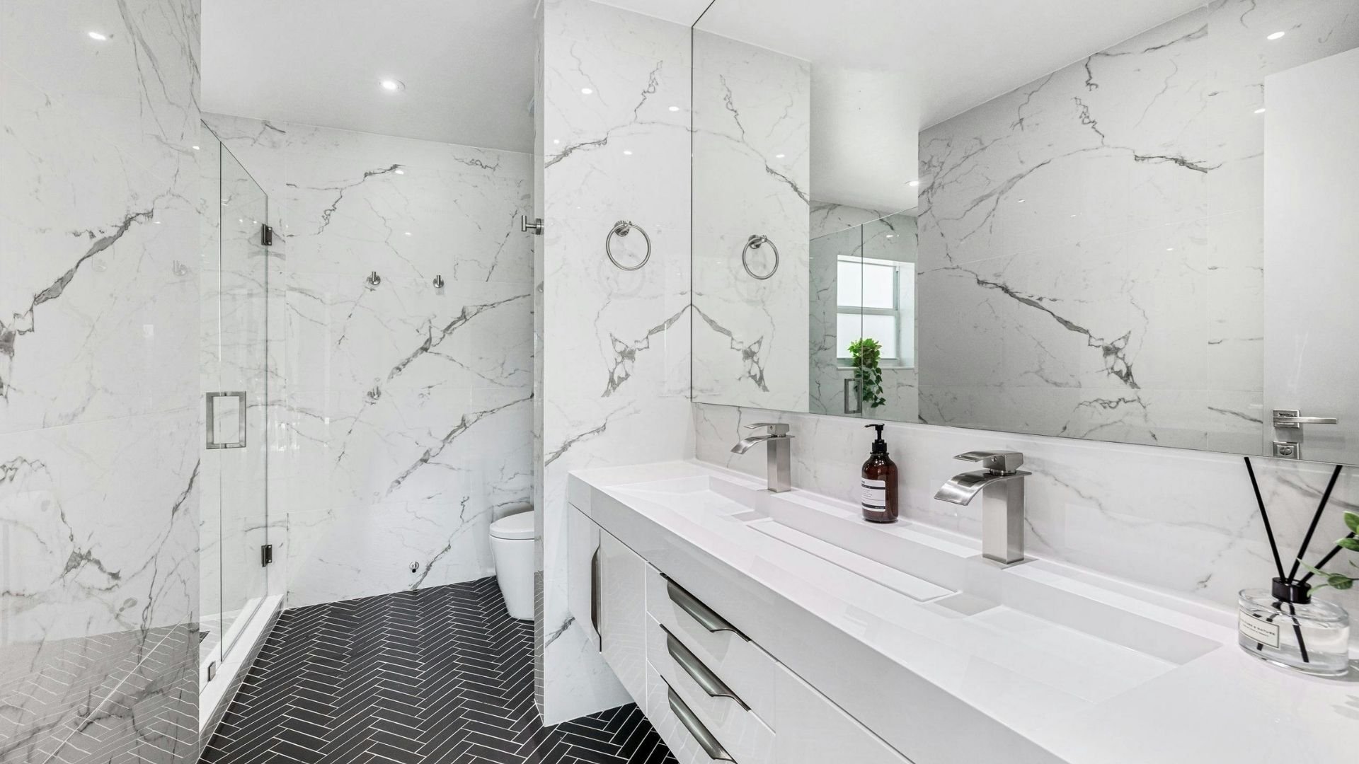 Modern bathroom with marble walls, double sink vanity, glass shower enclosure, and herringbone floor.