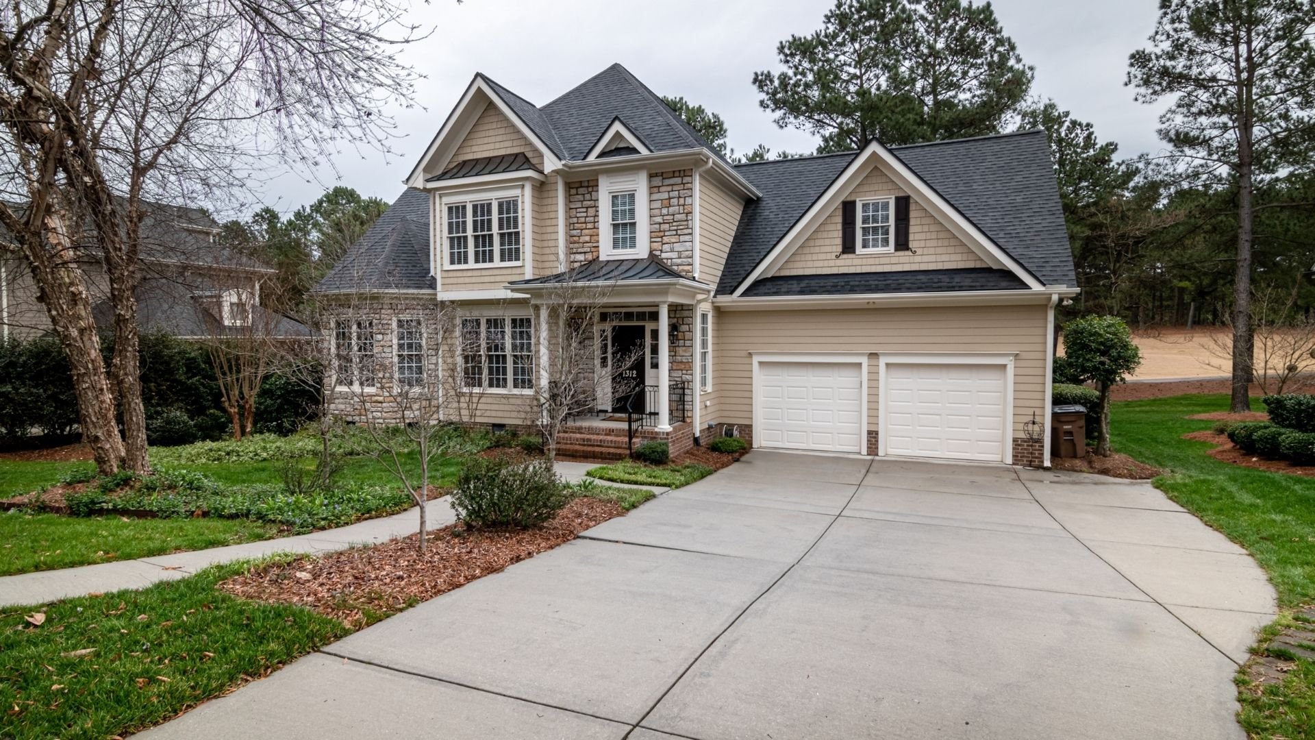 Two-story beige house with dark roof, white garage, and manicured landscaping on tree-lined suburban lot.