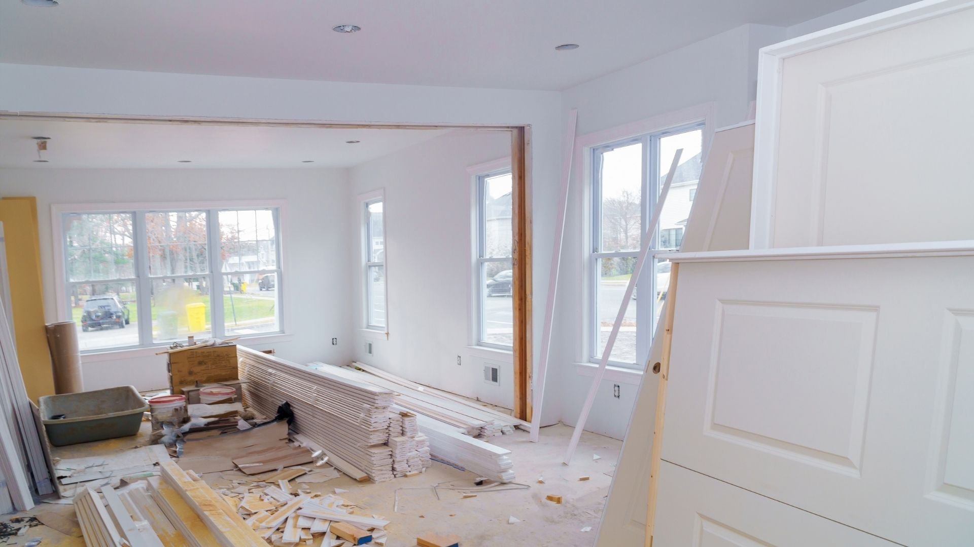 Spacious interior room under construction with white drywall, multiple windows, and building materials scattered throughout.